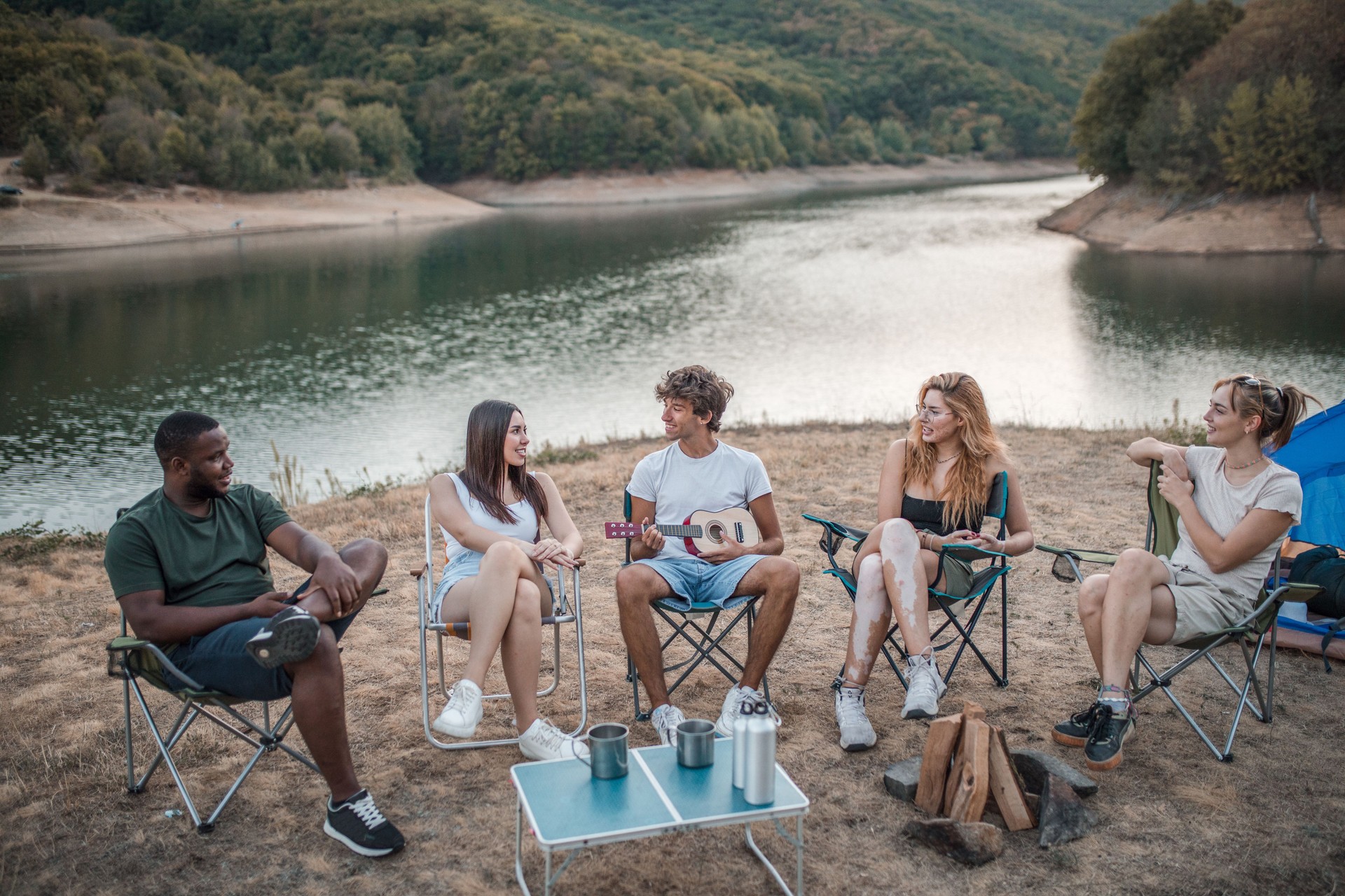 Handsome young man playing ukulele while camping with friends. They sit on the shore of the lake and hang out