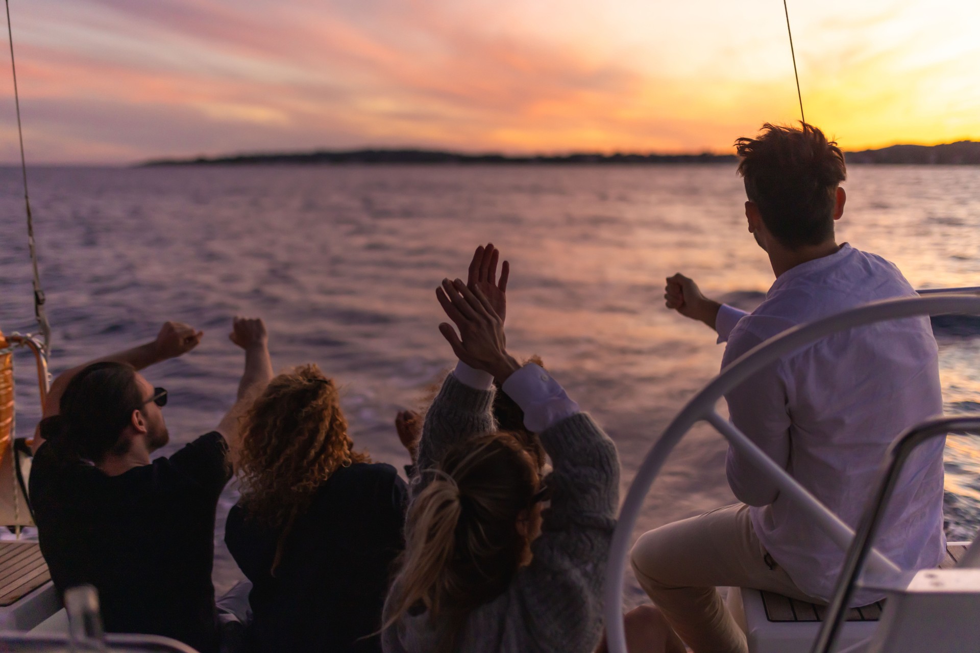 Group of friends sit on the back boat deck of a sailboat to watch the sunset, cheerful mood